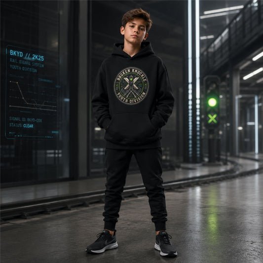 A young man wearing a A black pullover hoodie displayed on a plain white background, featuring a circular chest graphic with the text "Broken Knuckle Youth Division" wrapped around a technical design of a green railroad signal light, a crossing sign, and train tracks and matching black joggers stands in a futuristic industrial corridor.