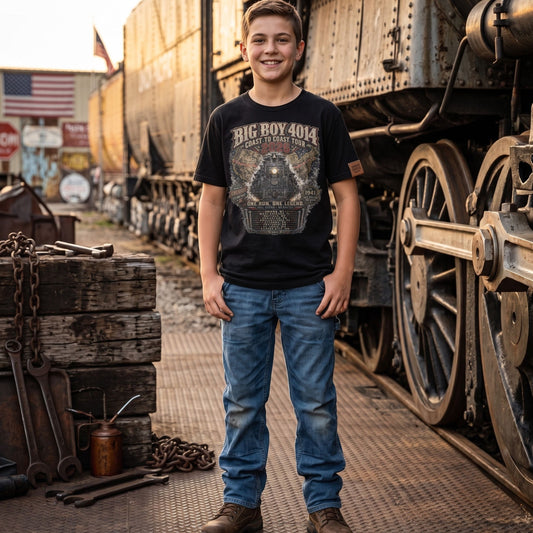 A boy wearing a Youth black t-shirt featuring a highly detailed vintage graphic of the Big Boy 4014 steam locomotive facing forward. Behind the train is a map of the United States showing the 2026 tour route. Text reads "BIG BOY 4014", "COAST TO COAST TOUR 2026", "ONE RUN. ONE LEGEND.", and "FULL STEAM - NO BRAKES". The graphic includes badges for "America's Greatest Steam Tour" and "Est 1941", along with a roster of the nine tour stops.