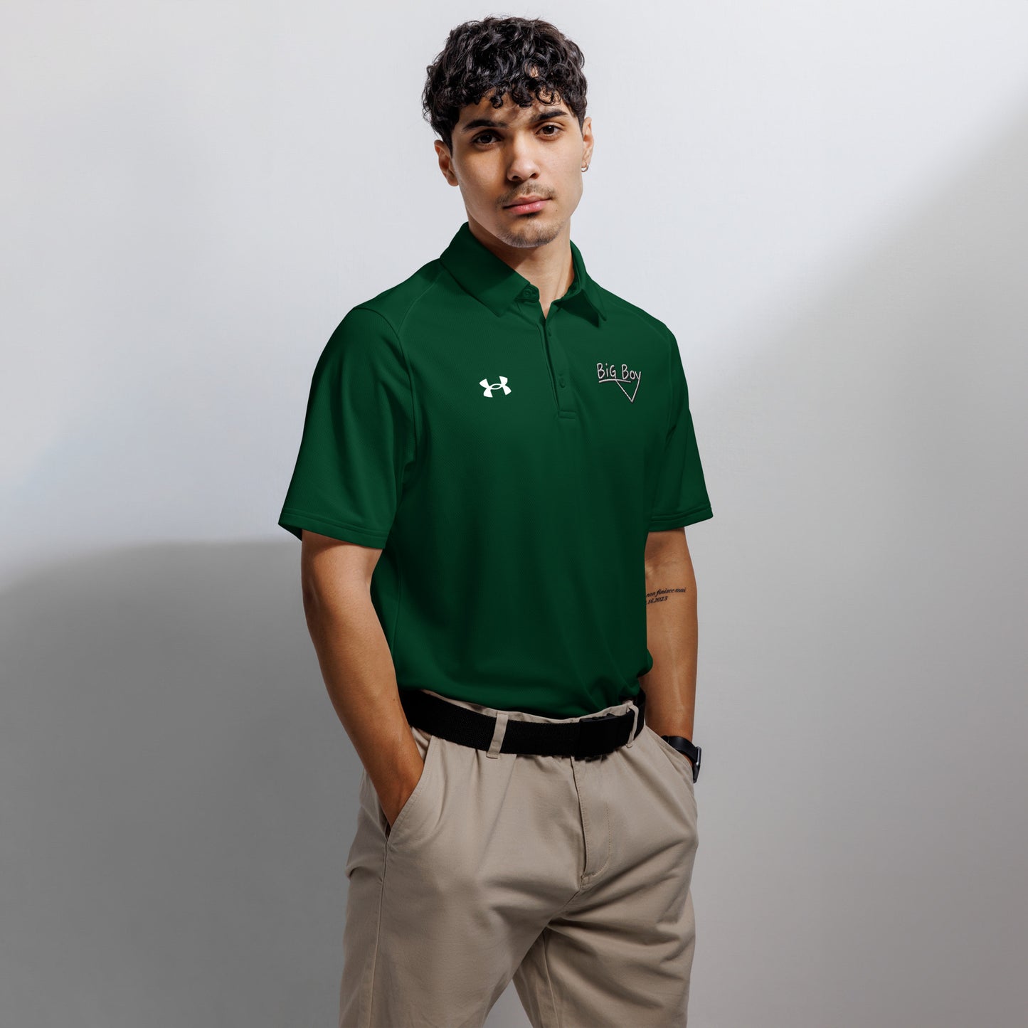 Forest green Under Armour men's polo shirt featuring the classic white chalk-style "Big Boy" script logo on the left chest. A white Under Armour logo is displayed on the right chest. The shirt has a classic collar and a three-button placket.