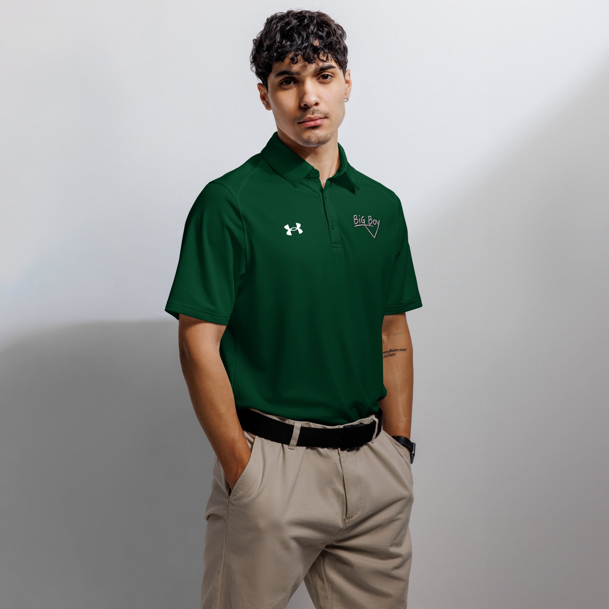 Forest green Under Armour men's polo shirt featuring the classic white chalk-style "Big Boy" script logo on the left chest. A white Under Armour logo is displayed on the right chest. The shirt has a classic collar and a three-button placket.