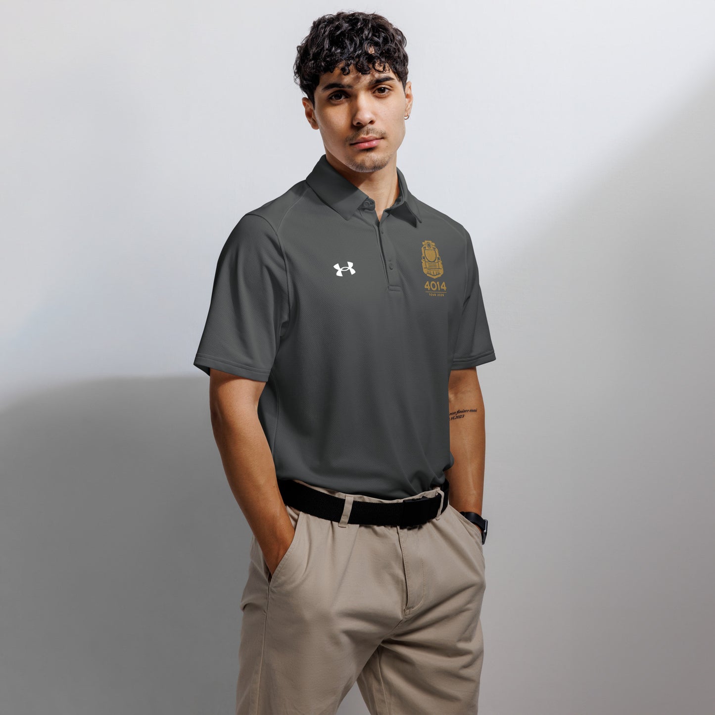 Gray Under Armour men's polo shirt featuring a gold graphic of a front-facing steam locomotive above the text "4014 TOUR 2026" on the left chest. A white Under Armour logo is displayed on the right chest. The shirt has a classic collar and a three-button placket.