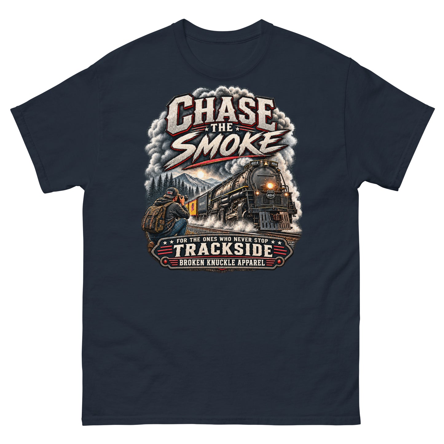 Navy classic t-shirt featuring a graphic of a photographer with a backpack kneeling trackside, taking a picture of a large steam locomotive rolling through a forest landscape under a setting sun. Massive plumes of smoke billow into the sky. Bold distressed text at the top reads "CHASE THE SMOKE". A vintage banner at the bottom reads "FOR THE ONES WHO NEVER STOP", "TRACKSIDE", and "BROKEN KNUCKLE APPAREL".