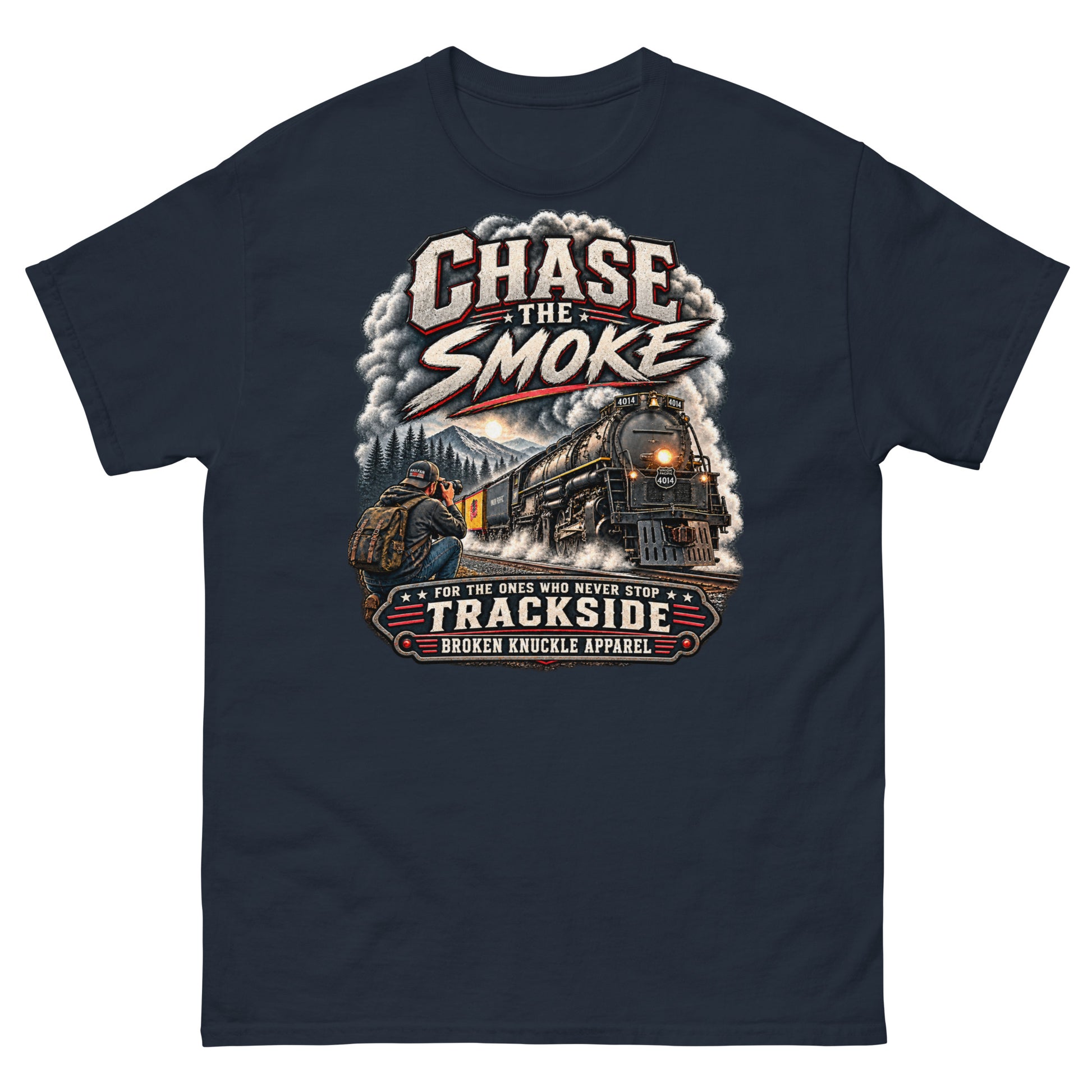 Navy classic t-shirt featuring a graphic of a photographer with a backpack kneeling trackside, taking a picture of a large steam locomotive rolling through a forest landscape under a setting sun. Massive plumes of smoke billow into the sky. Bold distressed text at the top reads "CHASE THE SMOKE". A vintage banner at the bottom reads "FOR THE ONES WHO NEVER STOP", "TRACKSIDE", and "BROKEN KNUCKLE APPAREL".