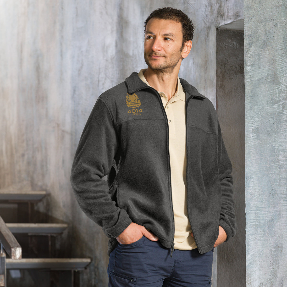 A man wearing a dark gray zip-up fleece jacket with a gold embroidered logo and the number '4014' on the left chest, layered over a beige polo shirt and navy pants, standing in a textured gray industrial setting.