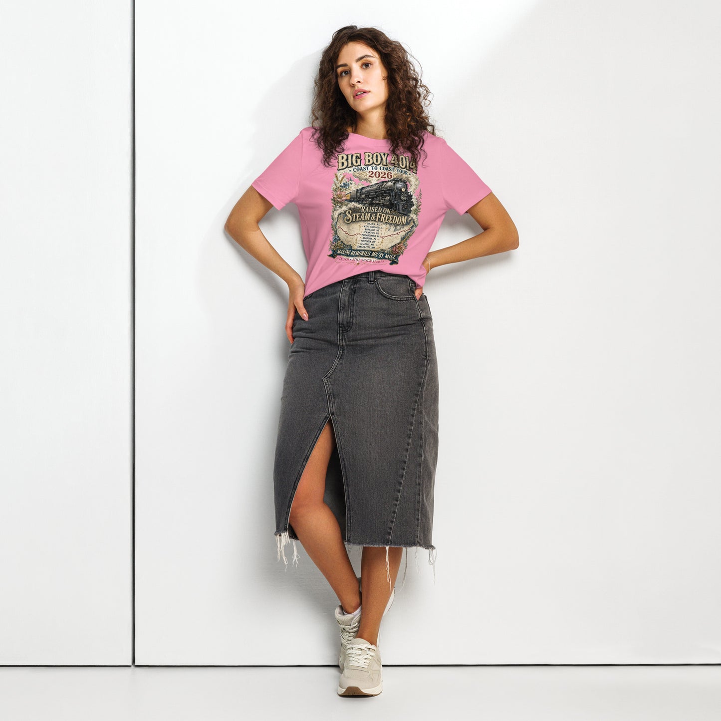 Woman wearing a pink t-shirt featuring a vintage illustration of the Big Boy 4014 steam locomotive surrounded by floral accents, a tour map, and text reading "BIG BOY 4014", "COAST TO COAST TOUR 2026", "RAISED ON STEAM & FREEDOM", and "MAKING MEMORIES MILE BY MILE".