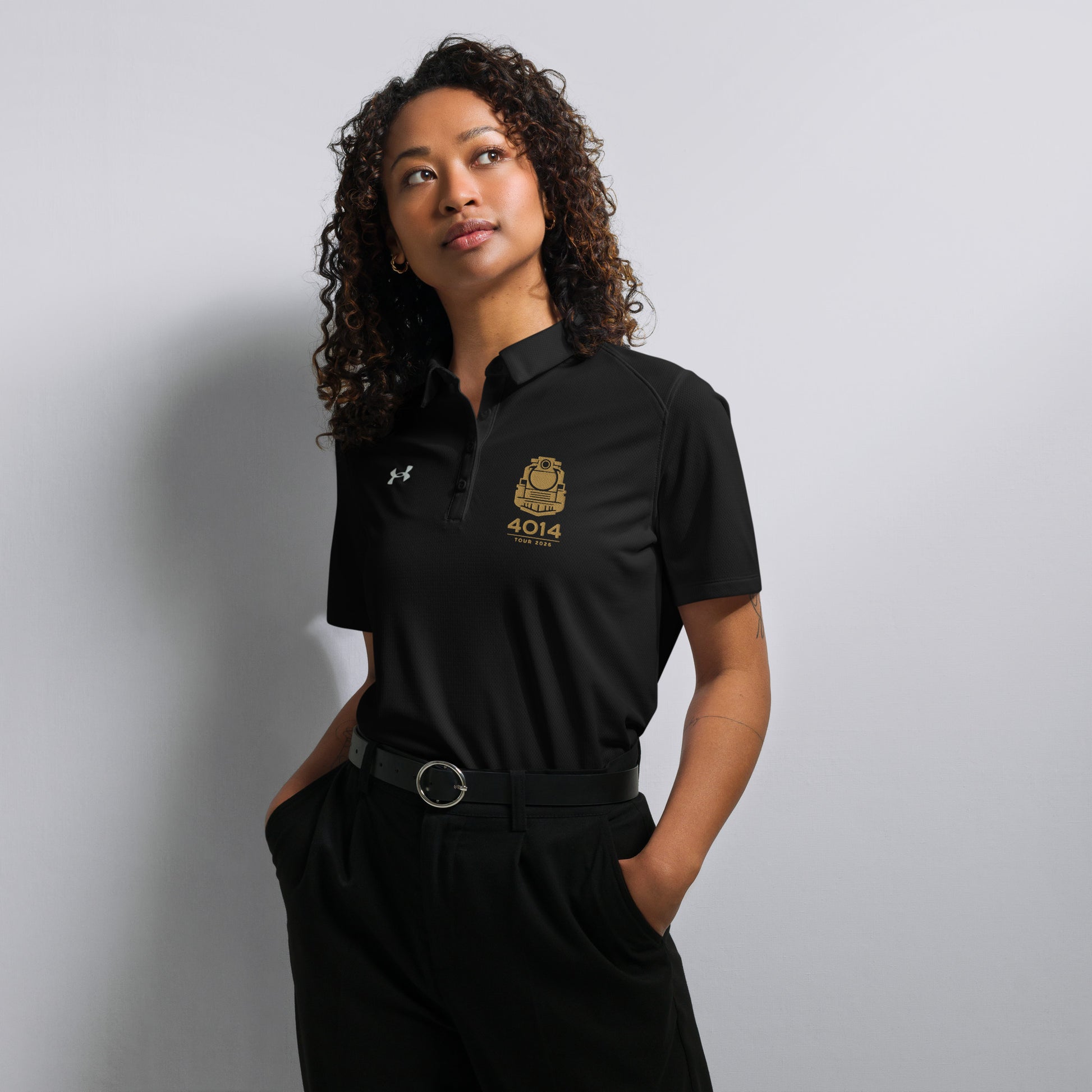 Black Under Armour women's polo shirt featuring a gold graphic of a front-facing steam locomotive above the text "4014 TOUR 2026" on the left chest. A white Under Armour logo is displayed on the right chest. The shirt has a classic collar and a multi-button placket tailored for a women's fit.