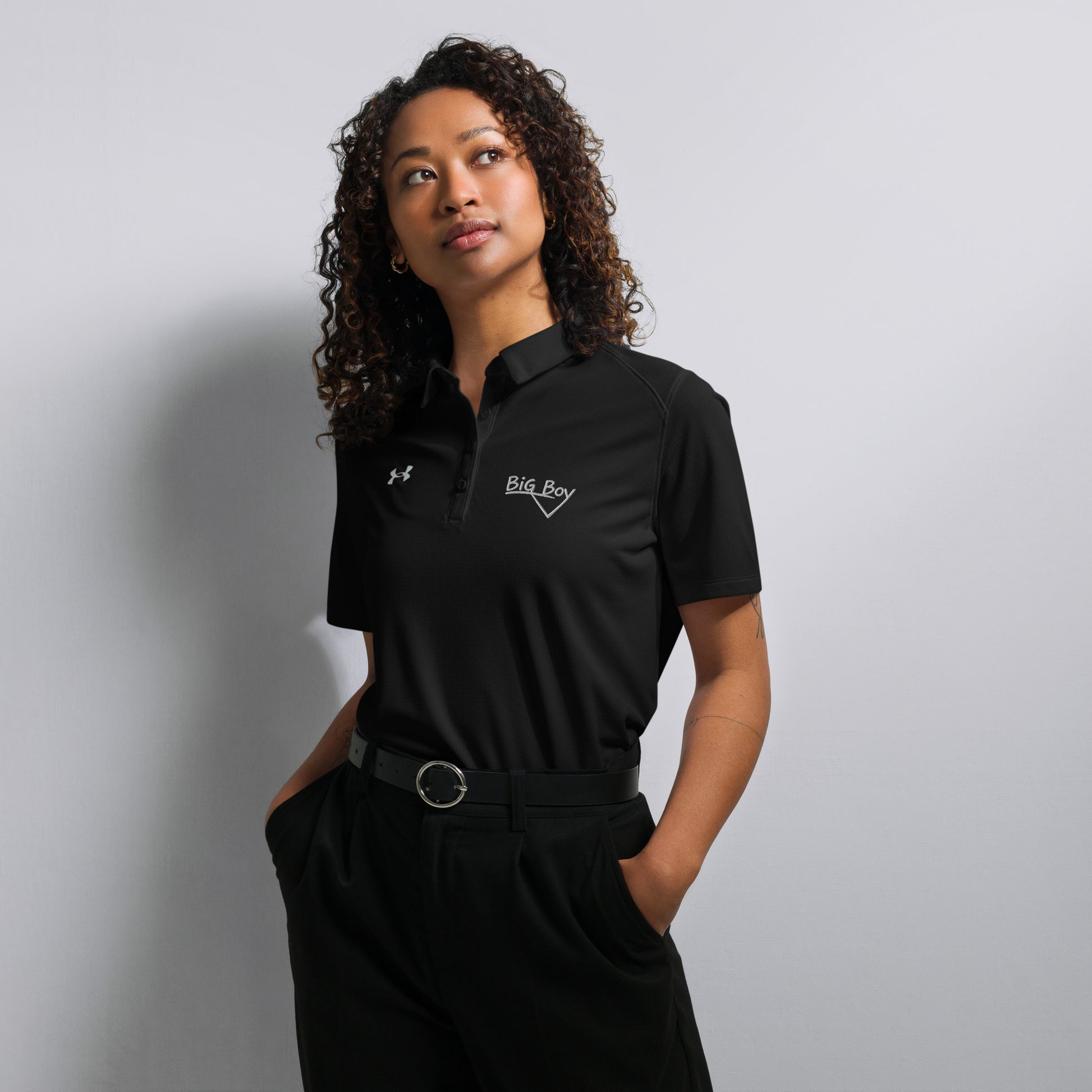 Black Under Armour women's polo shirt featuring the classic white chalk-style "Big Boy" script logo on the left chest. A white Under Armour logo is displayed on the right chest. The shirt has a classic collar and a multi-button placket tailored for a women's fit.