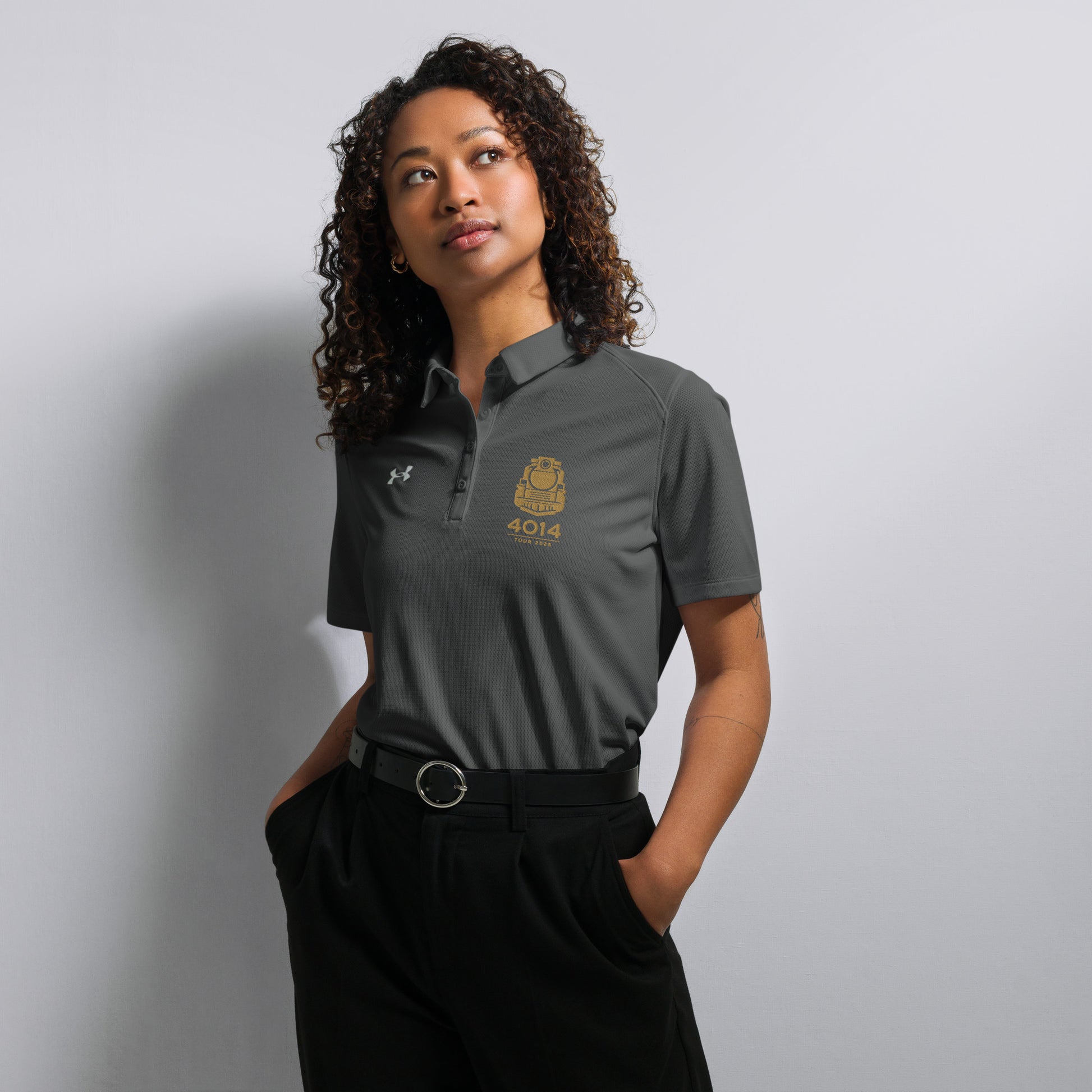 Gray Under Armour women's polo shirt featuring a gold graphic of a front-facing steam locomotive above the text "4014 TOUR 2026" on the left chest. A white Under Armour logo is displayed on the right chest. The shirt has a classic collar and a multi-button placket tailored for a women's fit.